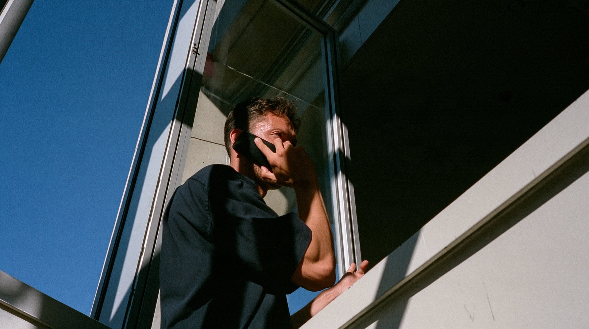 A man on a phone call at an open window against a deep blue sky