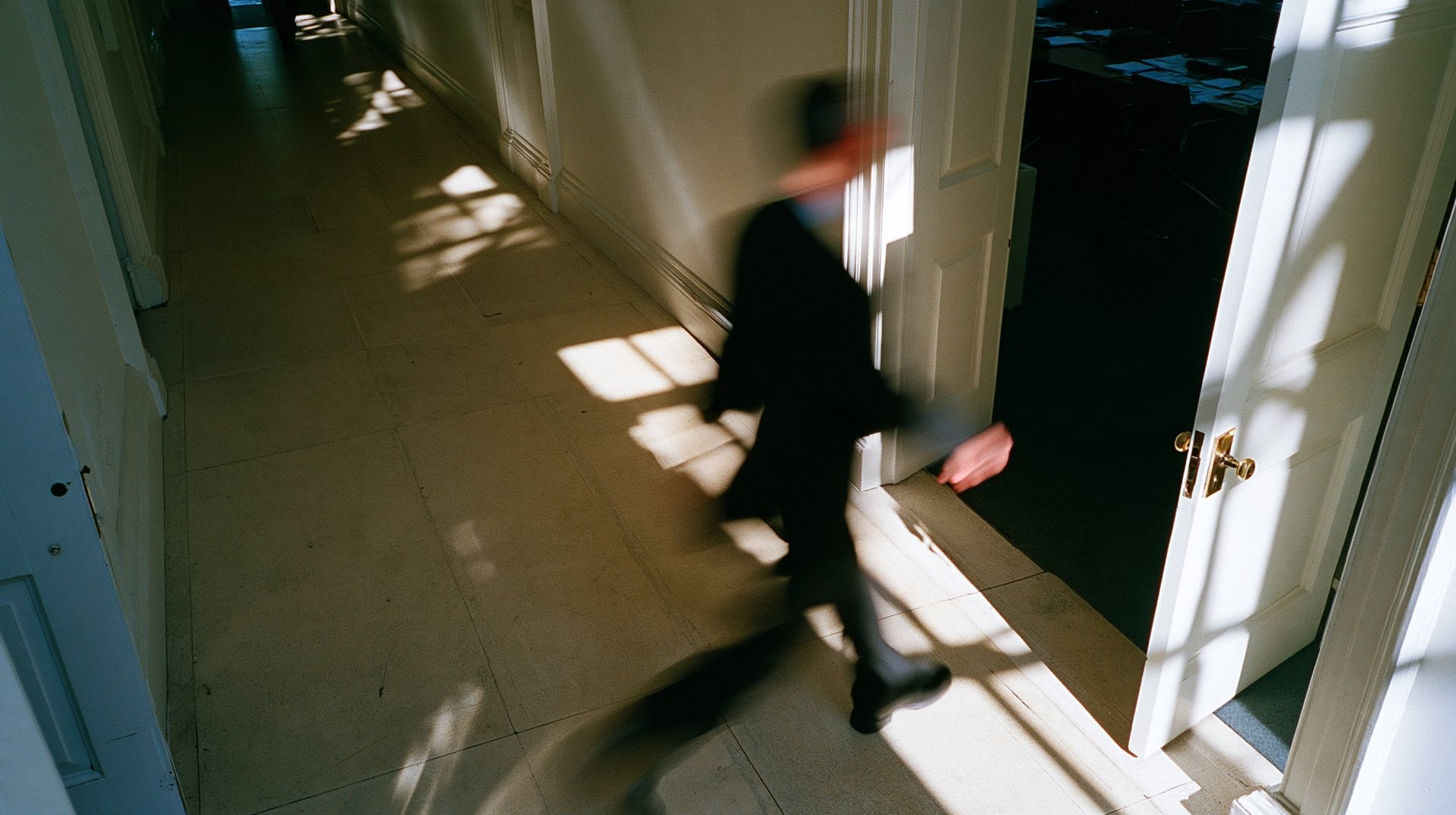 A figure in a dark suit striding through a doorway, blurred, in raking afternoon light
