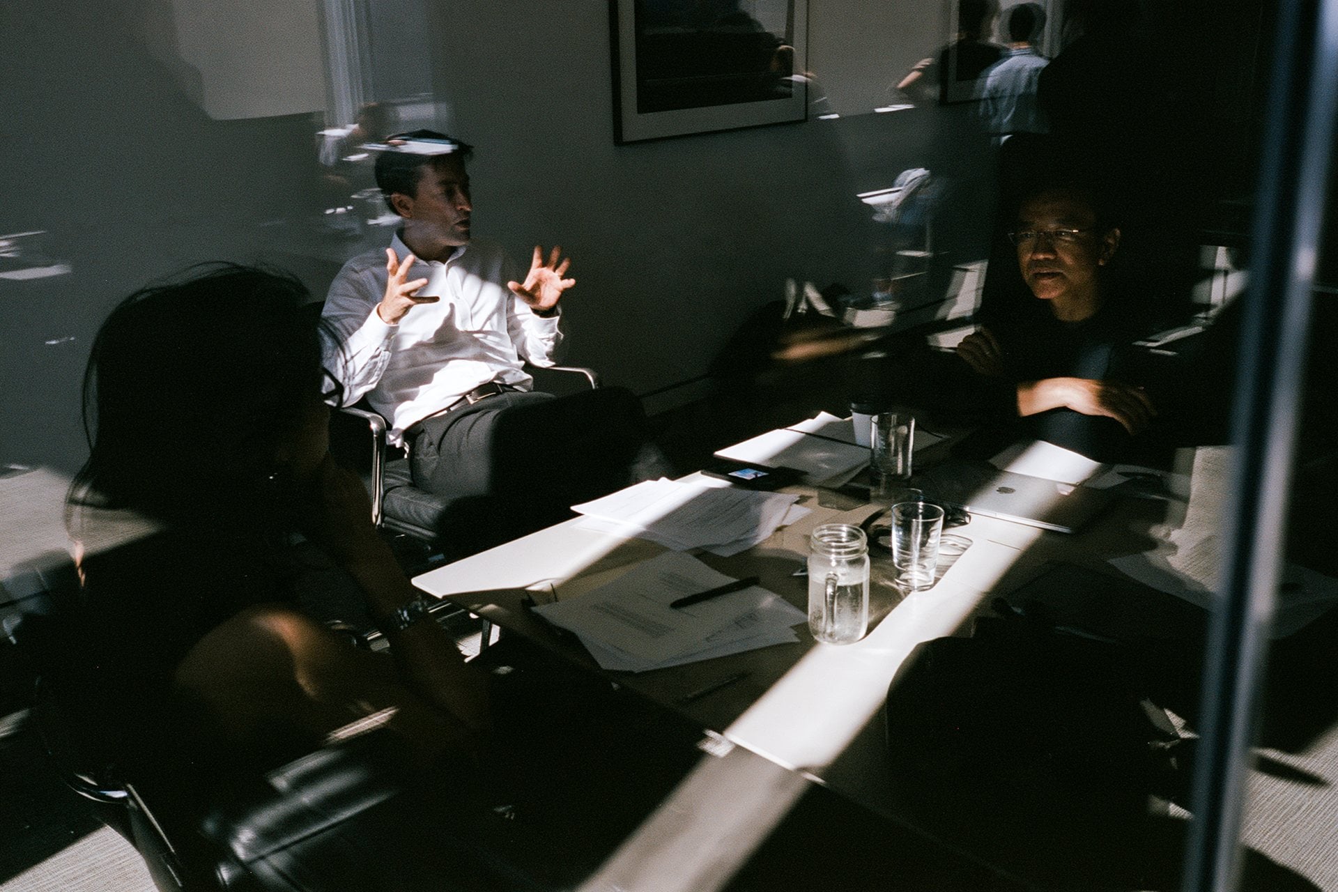 A meeting seen through glass, a man in a white shirt explaining across a sunlit table