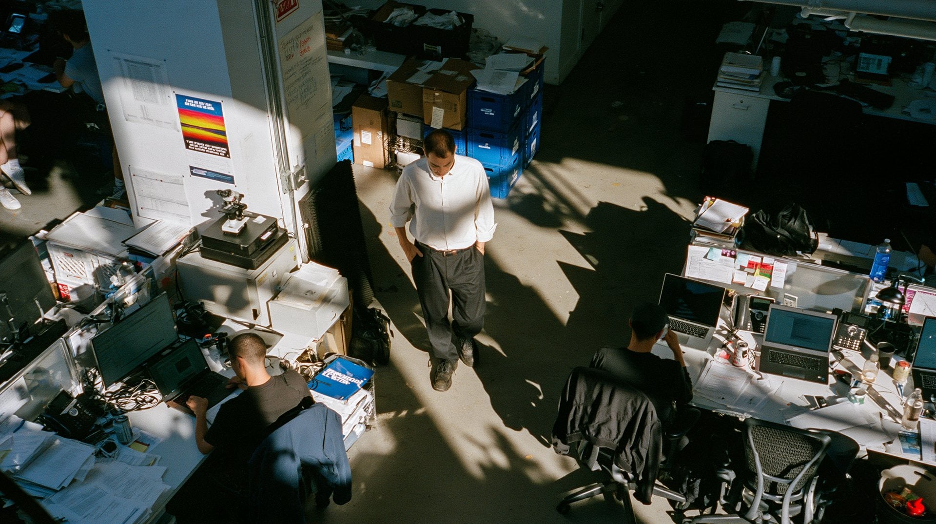 Looking down on a busy studio floor, a man in a white shirt walking between desks