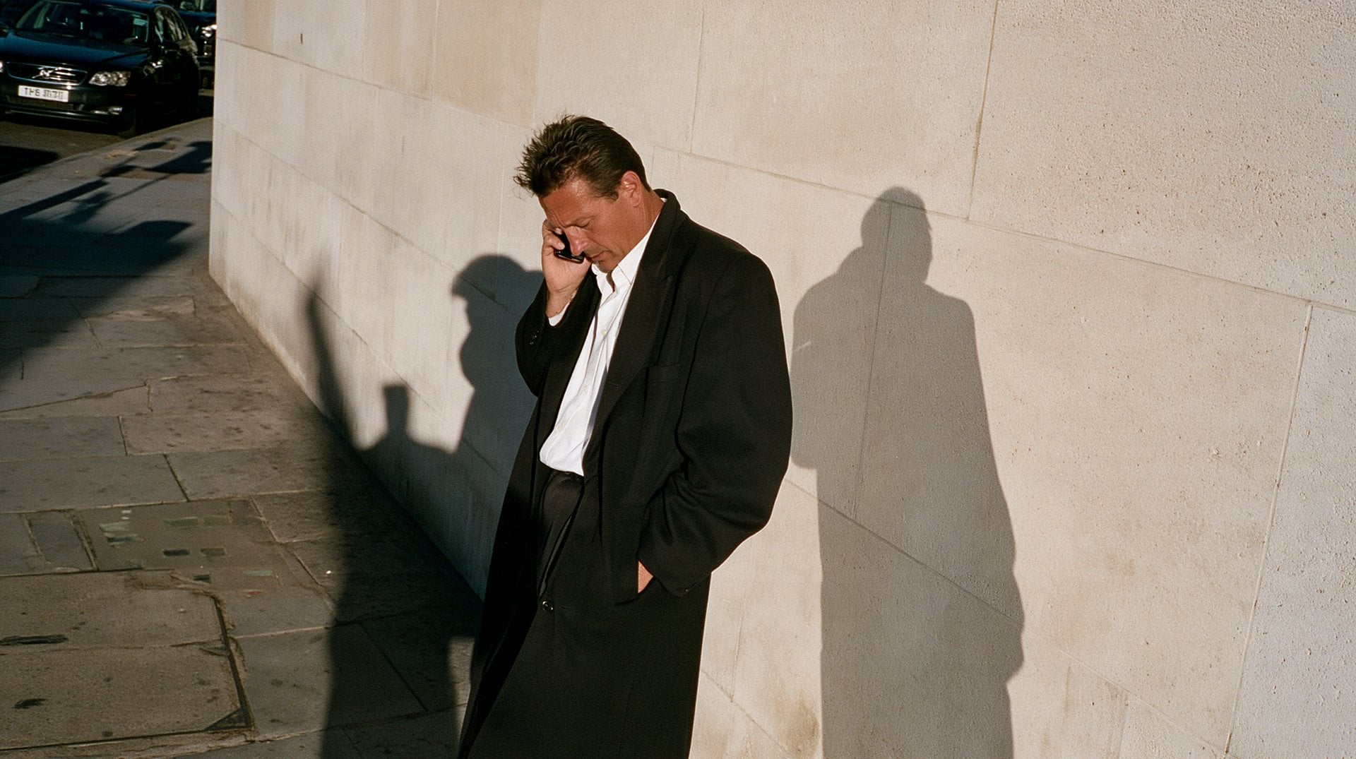 A man in a long coat on a phone call against a pale stone wall, his shadow beside him