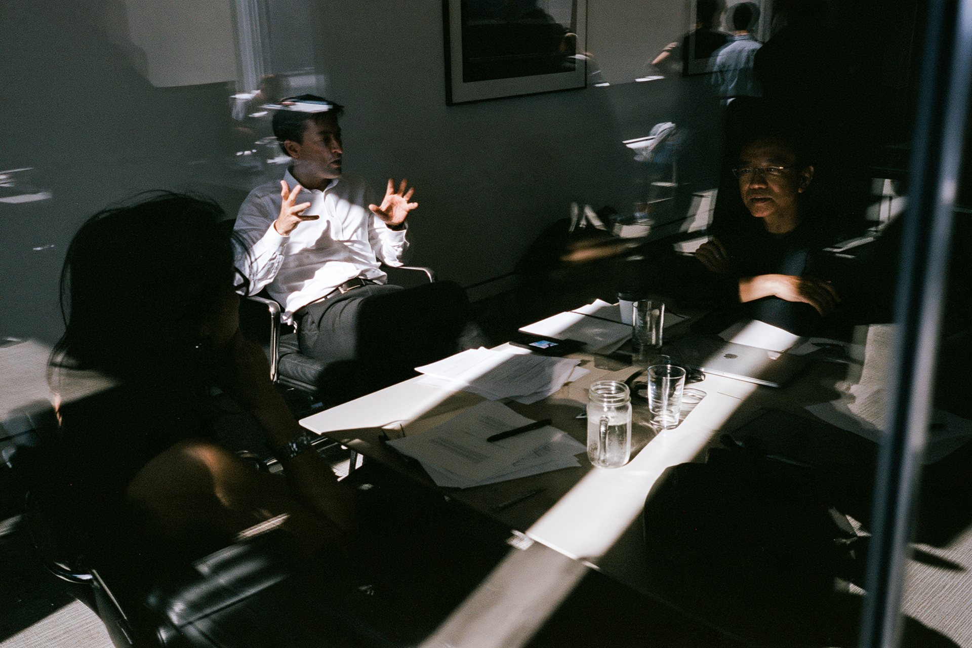 A meeting seen through glass, a man in a white shirt explaining across a sunlit table